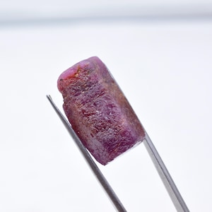 May include: A raw, uncut ruby gemstone held by tweezers. The ruby displays a deep, rich red-purple color with a rough, textured surface. The gemstone is rectangular in shape and appears to be a natural, unpolished specimen.