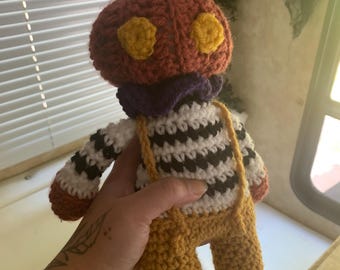 May include: A handmade crochet doll with a pumpkin-shaped head, yellow eyes, and a purple scarf. The doll wears a black and white striped shirt, yellow overalls, and brown shoes. The doll is held in a person's hand.