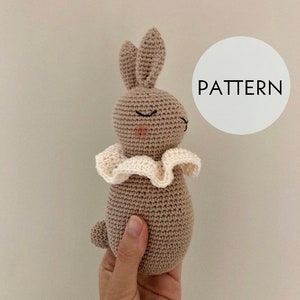 Può includere: Un giocattolo a forma di coniglietto all'uncinetto nei toni del beige e del crema. Il coniglietto ha gli occhi chiusi, guance rosa e un colletto a balze color crema. La parola "PATTERN" è in un cerchio bianco sullo sfondo.