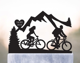 May include: Black silhouette cake topper of a couple riding bicycles in front of a mountain range with a heart and the date 14.03.2019.