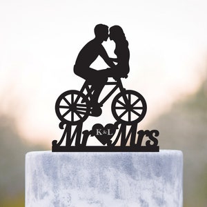 May include: Black silhouette of a couple kissing while riding a bicycle, with the words "Mr & Mrs" and the initials "K & L" in a heart below. This is a wedding cake topper.
