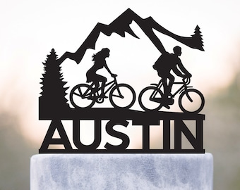 May include: Black cake topper featuring a mountain scene with two cyclists and a pine tree. The word "AUSTIN" is displayed below the cyclists. The topper is placed on a white cake with a bow and a decorative jewel.