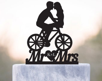 May include: Black silhouette of a couple kissing while riding a bicycle, with the words "Mr & Mrs" and the initials "K & L" in a heart below. This is a wedding cake topper.