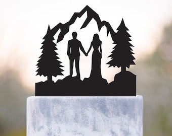 May include: Black silhouette cake topper of a couple holding hands in front of a mountain range with two pine trees on either side.