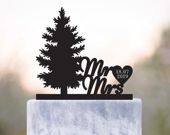 May include: A black wooden cake topper with a pine tree silhouette and the cursive words "Mr Mrs". The date "18.07 2019" is inside a heart. The topper sits on a multi-tiered white cake, decorated with a bow and a jewel.