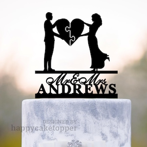Op de afbeelding: Zwarte silhouet van een stel dat een hartvormig puzzelstuk vasthoudt, met de tekst "Mr. & Mrs. ANDREWS" eronder. Het stel staat op een witte taart met een grijze marmeren laag.