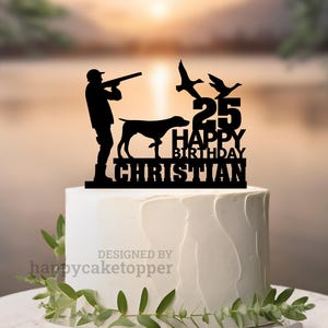 May include: A black cake topper with a hunting theme, featuring a silhouette of a hunter, dog, and flying ducks. The topper includes the text "25 HAPPY BIRTHDAY CHRISTIAN". The cake is white with a simple design, and greenery is placed around the base.