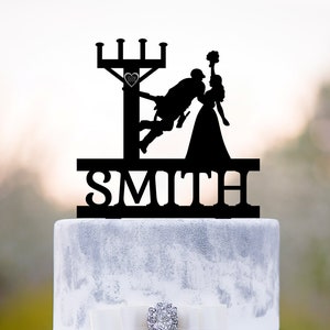 Op de afbeelding: Zwarte silhouet-taarttopper van een bruid en bruidegom. De bruidegom klimt op een elektriciteitspaal en de bruid houdt een boeket bloemen vast. De tekst "SMITH" staat onder het paar.
