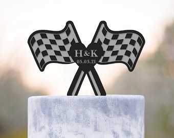Könnte beinhalten: Ein Tortenaufsatz mit schwarz-weiß karierten Flaggen, den Initialen "H&K" und dem Datum "05.03.21" in Herzform. Die Torte ist weiß mit silbernen Akzenten und einer dekorativen Schleife mit Strassbesatz.