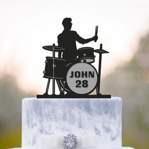 May include: Black silhouette of a drummer playing a drum set with the name "JOHN 28" on the bass drum. The cake topper is on top of a two-tiered wedding cake.