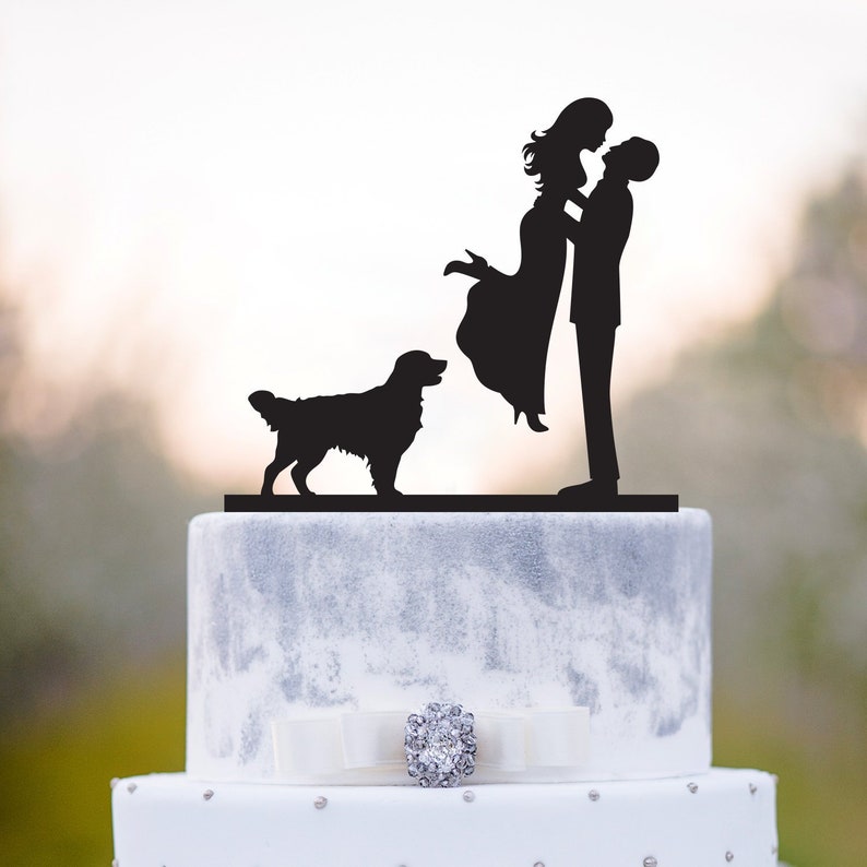 May include: Black silhouette cake topper of a couple kissing with a golden retriever dog standing in front of them. The topper is on a white and gray wedding cake.