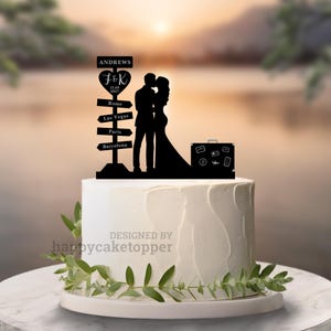 Puede incluir: Un adorno para tarta negro con la silueta de una pareja besándose, una maleta y un poste indicador con nombres y destinos. La tarta es blanca con glaseado texturizado y vegetación.