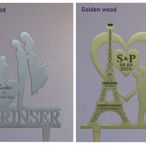 May include: Two wedding cake toppers, one silver and one gold. The silver topper features a couple kissing with a dog, a heart with the names "Nicolai & Veronika", and the word "LORINSER". The gold topper features a couple holding hands in front of the Eiffel Tower, a heart with the initials "S&P" and the date "08.08.2020".