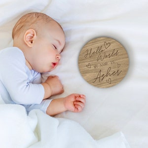 Può includere: Un cartello rotondo di legno con il testo "Hello World My Name Is Asher" e cuori intorno al testo. Il cartello è su una superficie bianca con un bambino che dorme sopra.
