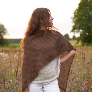 May include: A brown knitted poncho with a tassel, worn over a white top and white trousers. The person is standing in a field of wildflowers.
