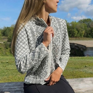 Può includere: Una giacca di maglia bianca con un motivo floreale nero e una cerniera. La giacca è indossata da una persona con lunghi capelli biondi.