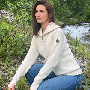 May include: A woman wearing a white sweater with a zipper and a small brown patch on the left side of the chest. She is wearing blue jeans and is squatting in a grassy area near a stream.