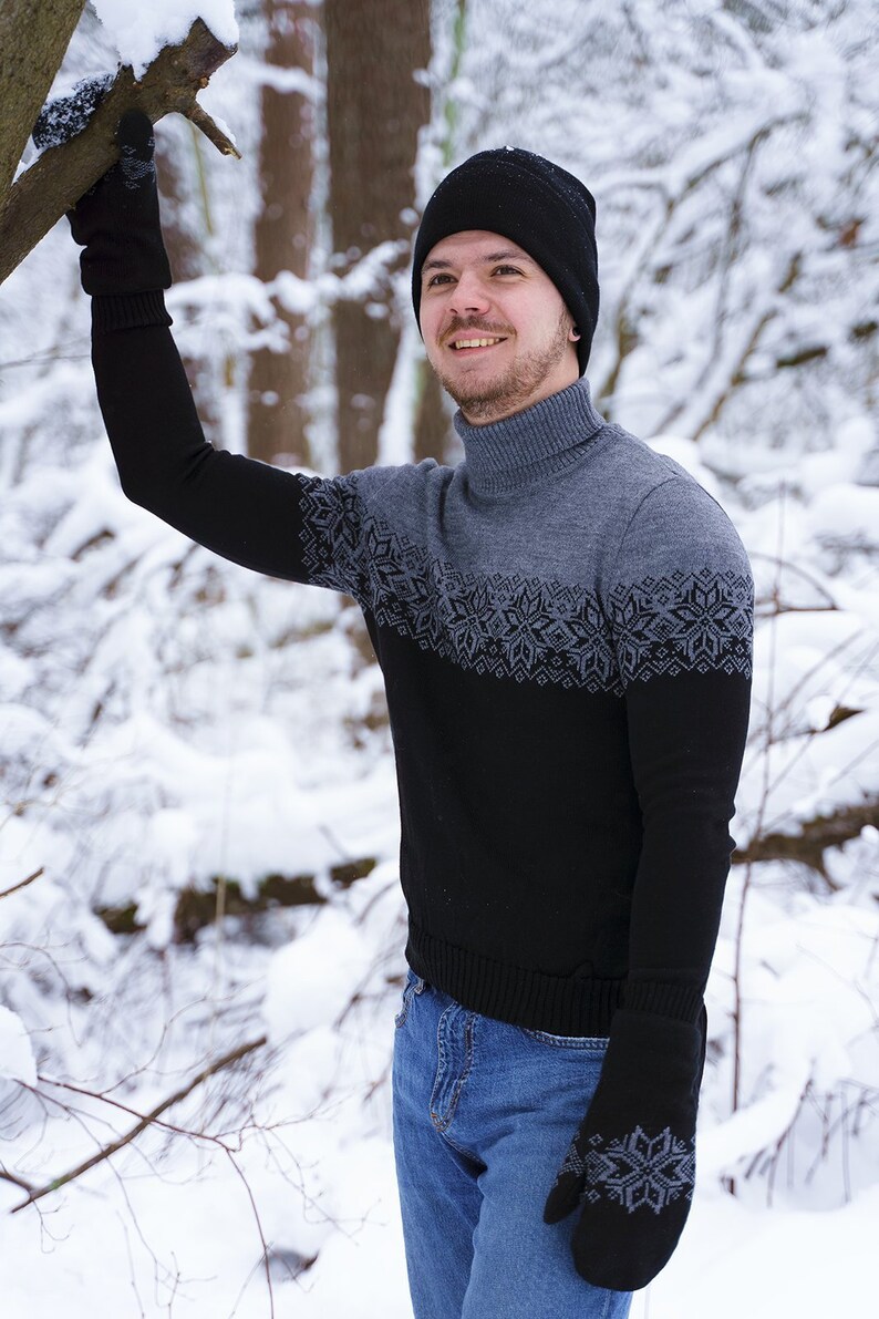 May include: A man wearing a black knit hat and a black and gray sweater with a snowflake pattern. He is wearing blue jeans and black gloves. The man is standing in a snowy forest.