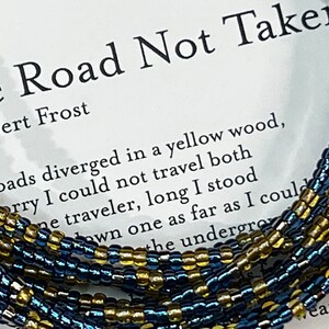 May include: A close-up of a beaded bracelet with blue and gold beads. The bracelet is on a white background with the text "The Road Not Taken" by Robert Frost.