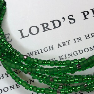 May include: A green beaded bracelet with silver accents. The bracelet is made of multiple strands of beads and is wrapped around a book with the text "LORD'S PRAYER" visible.