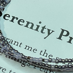 May include: A close-up of a beaded bracelet with a silver and purple color scheme. The bracelet is lying on a white card with the text "Serenity Prayer" printed on it.