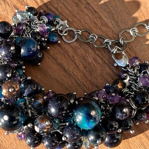 May include: A silver chain bracelet with a cluster of dark blue, black, and purple beads. The beads are of various sizes and shapes, and some have a sparkly finish.