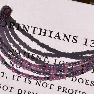 May include: A multi-strand bracelet made of small purple and silver beads. The bracelet is on a white background with the text "1 Corinthians 13:  Patient, Love Does Not..." printed on it.