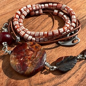 May include: A multi-strand necklace with brown leather cord and red and white striped beads. The necklace features a large round red and brown stone pendant, a smaller red stone, and a smaller brown stone pendant.