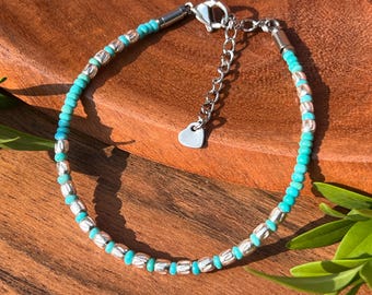 May include: A turquoise and silver beaded bracelet. The bracelet features small, round turquoise beads interspersed with silver-colored, oblong beads. It has a silver chain extender and a small heart-shaped charm.