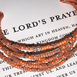 May include: A multi-strand beaded bracelet with peach-colored beads and silver accents. The bracelet is on a white surface with the text "The Lord's Prayer" printed on it.