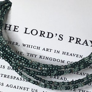 May include: A green beaded bracelet with gold accents. The bracelet is wrapped around a piece of paper with the text "The Lord's Prayer" printed on it.