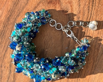 May include: A blue and green glass bead bracelet with a silver clasp and a silver charm. The bracelet is made of a variety of different sized beads, some of which are iridescent.