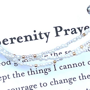 May include: A delicate white beaded bracelet with gold accents, displayed on a white card. The card features the text "Serenity Prayer" and a portion of the prayer. The bracelet is made of small, round beads.