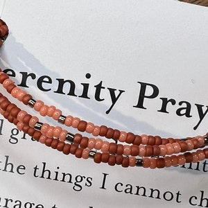 May include: A multi-strand beaded bracelet with brown and peach colored beads. The bracelet is on a white card with the text "Serenity Prayer" printed on it.