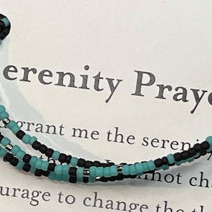 May include: A beaded bracelet with black and turquoise beads. The bracelet is on a white card with the text "Serenity Prayer" and a portion of the prayer.