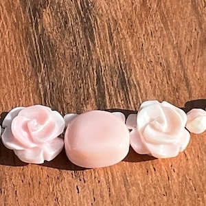 May include: A pink shell necklace with carved rose beads and a larger oval bead. The necklace is on a wooden surface.