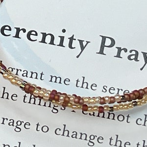 May include: A beaded bracelet with brown and gold beads, with the text "Serenity Prayer" and a portion of the prayer text visible in the background.