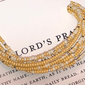 May include: A multi-strand beaded bracelet with alternating cream and gold beads. The bracelet is lying on a white card with the text "The Lord's Prayer" printed on it.