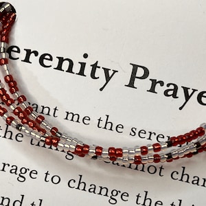 May include: A red, white, and black beaded bracelet with the text "Serenity Prayer" and the words "Grant me the serenity to accept the things I cannot change; courage to change the things I can; and the wisdom to know the difference."