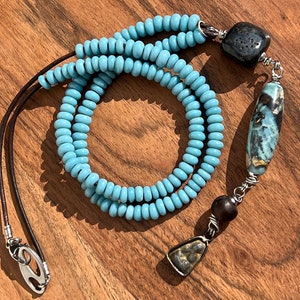 May include: A long blue beaded necklace with a brown leather cord and three silver metal charms. The charms are a dark brown stone with holes, a light blue and gray stone, and a small silver charm with a dark gray stone.