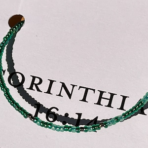May include: A green beaded bracelet with silver beads and the text "CORINTHIAN 16:14" on a white background.