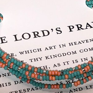 May include: A multi-strand bracelet made of teal and orange beads with silver accents. The bracelet is lying on a page of a book with the text "The Lord's Prayer" printed on it.