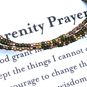 May include: Close-up of a beaded bracelet with three strands. The beads are primarily olive green with sections of gold and copper-colored beads. The text "Serenity Prayer" and part of the prayer are visible in the background.