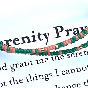 May include: A beaded bracelet with alternating green and peach-colored beads. The bracelet is displayed over a white card with the text "Serenity Prayer" and part of the prayer printed on it.