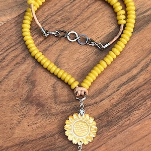 May include: A yellow beaded necklace with a sunflower pendant. The pendant is made of carved wood and has a leaf charm.