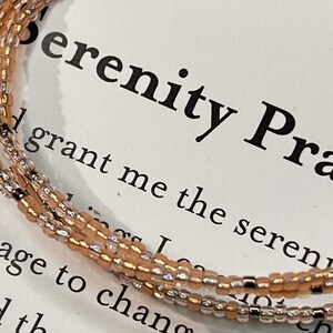 May include: A triple-strand beaded bracelet with a light brown and silver color scheme. The bracelet is on a card with the text "Serenity Prayer" and a portion of the prayer: "Grant me the serenity..."