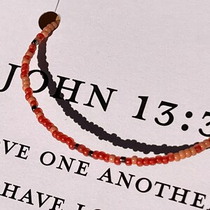 May include: A beaded bracelet with red and beige beads and black accents. The bracelet has the text "JOHN 13:34 LOVE ONE ANOTHER." printed on a white card.