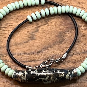 May include: A necklace with a black leather cord and light green beads. The necklace has a silver clasp and a pendant made of black and white stone.