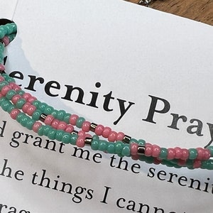 May include: A close-up of a beaded bracelet with pink and green beads. The bracelet is on a white card with the text "Serenity Prayer" and "God grant me the serenity to accept the things I cannot change."