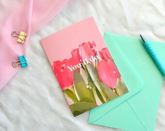 May include: A pink greeting card with a floral design of red tulips and the text "You'll do". The card is next to a light green envelope, a blue pen, and two binder clips. A pink fabric is draped in the background.